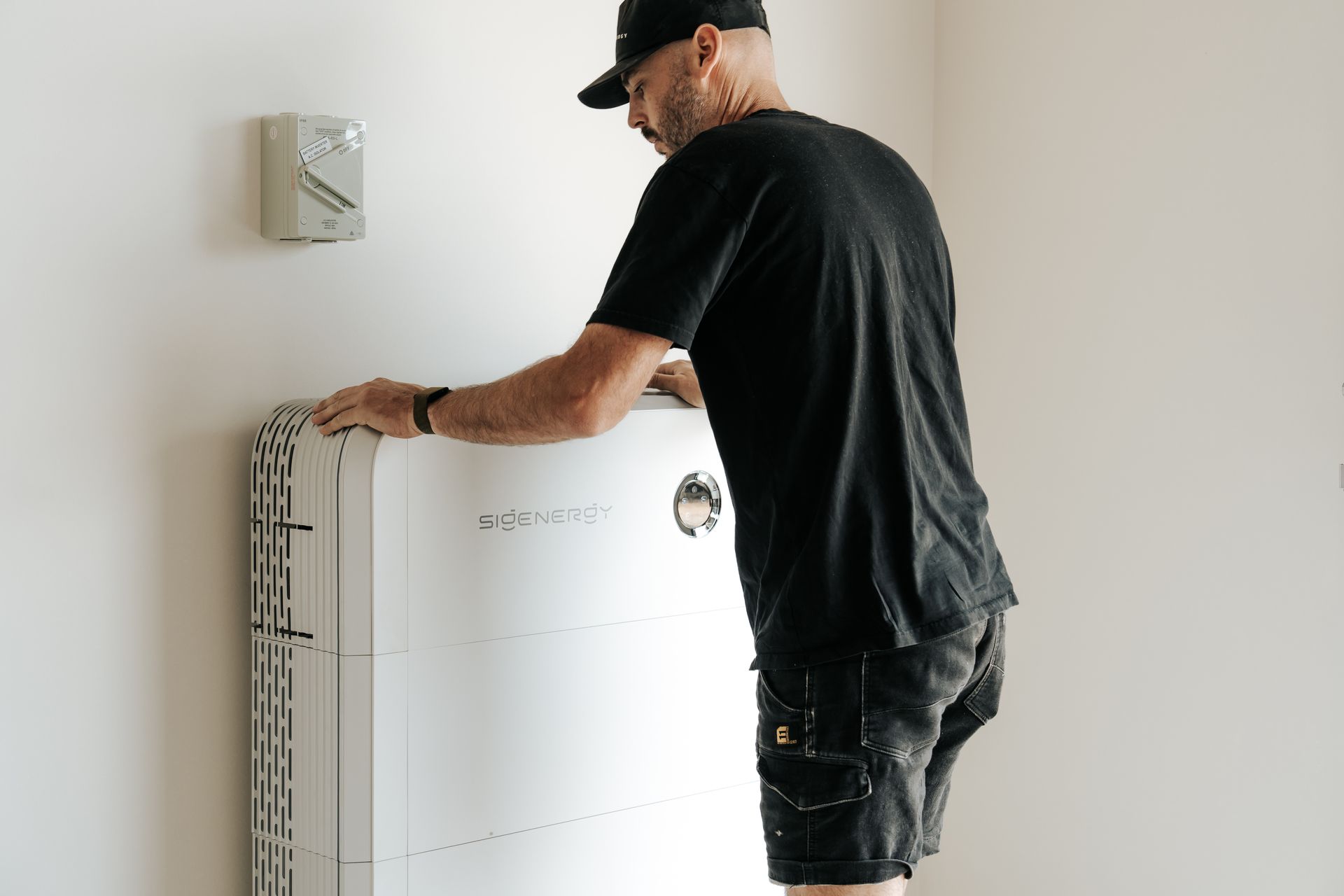 A person in a black shirt and shorts installs a white wall-mounted home battery unit next to a wall-mounted controller.