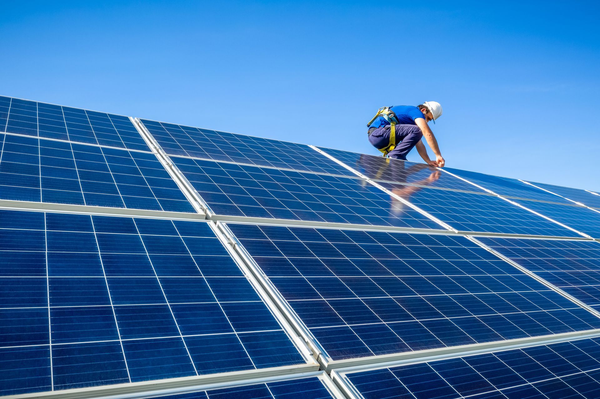 Person in safety harness installing solar panels on a roof against a blue sky.