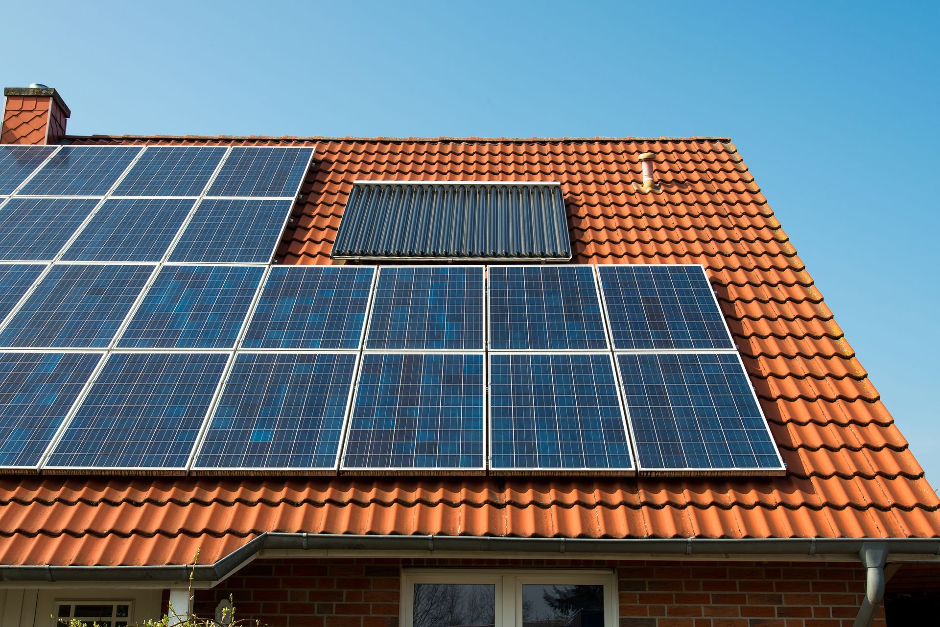 Solar panels and a solar thermal collector on a red-tiled roof against a clear blue sky.