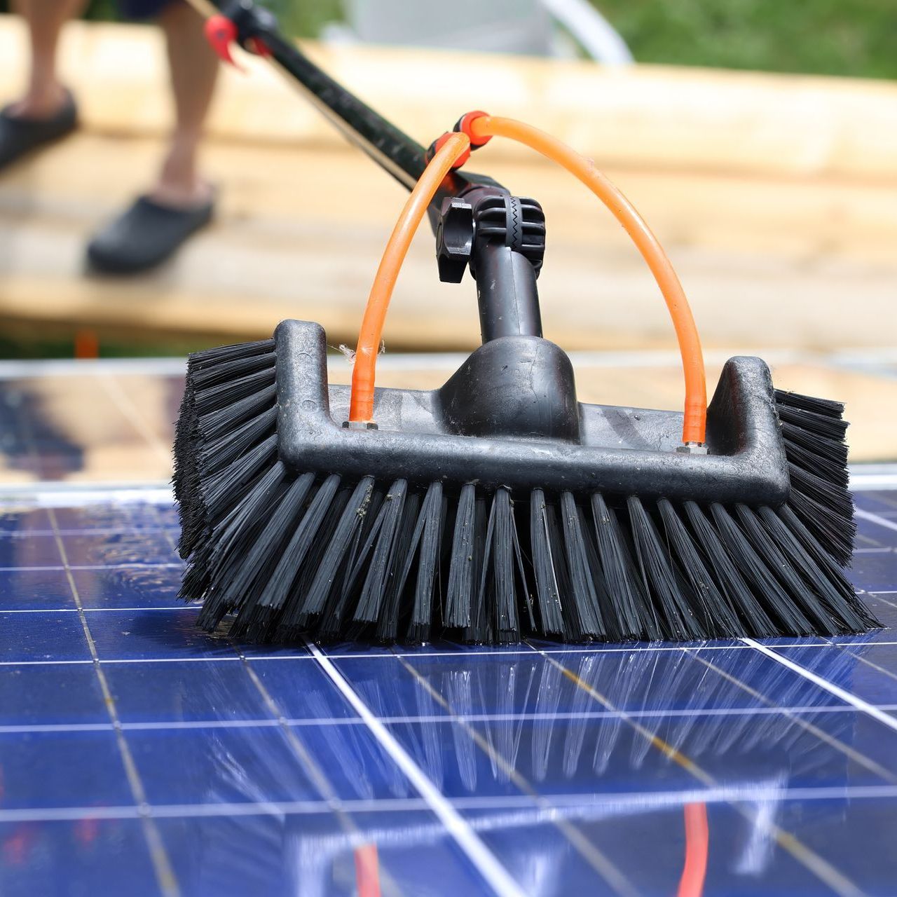 Solar panel being cleaned with a brush attached to a long handle; orange water tubes visible.