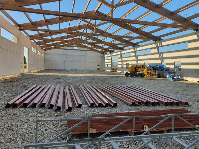 Un cantiere edile che mostra la struttura di un grande magazzino a cielo aperto, con travi d'acciaio disposte ordinatamente su un pavimento di ghiaia.