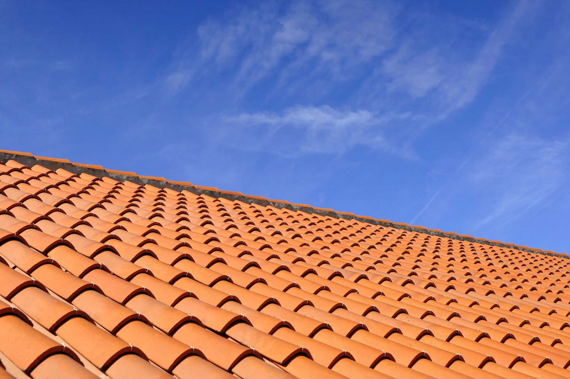 Una vista dal basso di un tetto di tegole in terracotta che si innalza verso l'alto, stagliandosi contro un cielo azzurro brillante punteggiato da nuvole leggere.