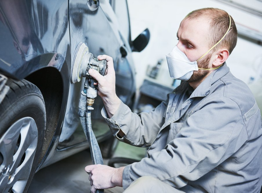Man using a sander on a car door, wearing a respirator mask, in a garage.