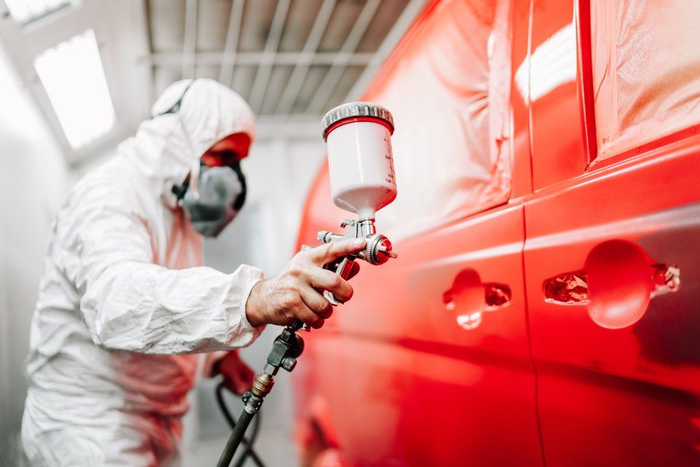 Person in protective suit sprays red paint on a vehicle in a paint booth.