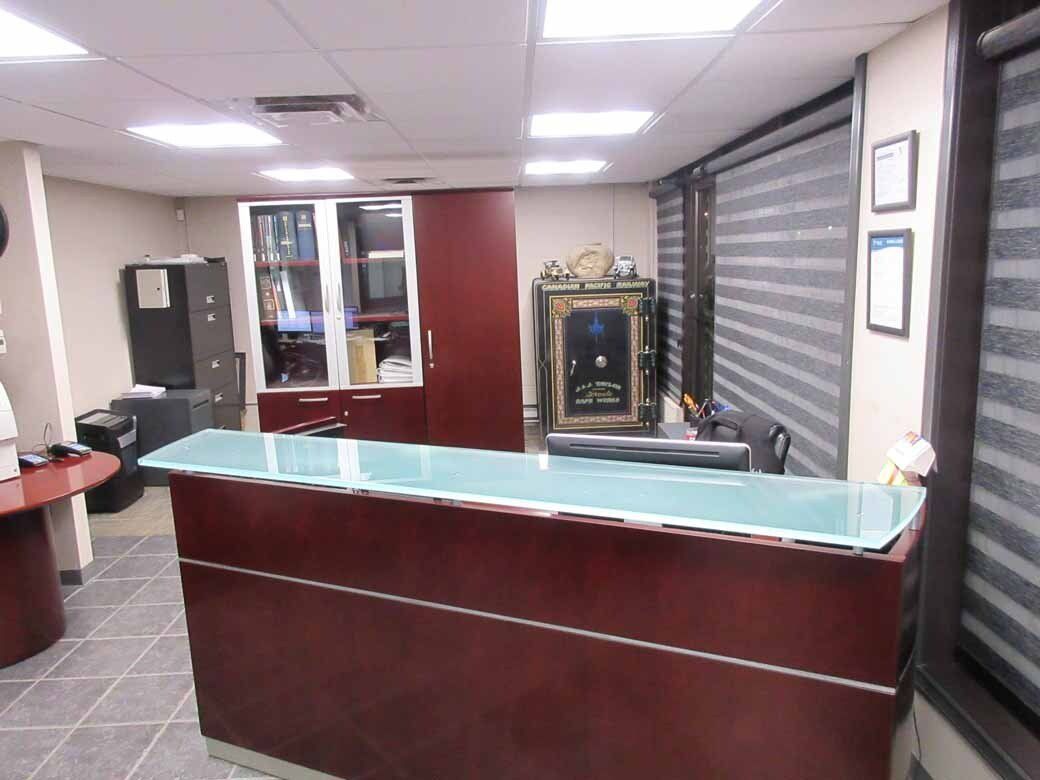 Reception desk in an office. Reddish-brown desk with glass top, cabinets, blinds, and overhead lighting.
