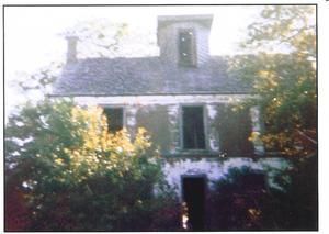 An abandoned, weathered two-story house with a small central dormer window, surrounded by overgrown trees and foliage.