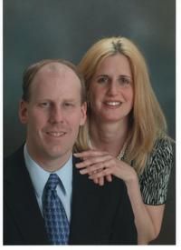 A professional portrait of a man in a suit and tie and a woman in a patterned blouse, posing closely against a grey backdrop.