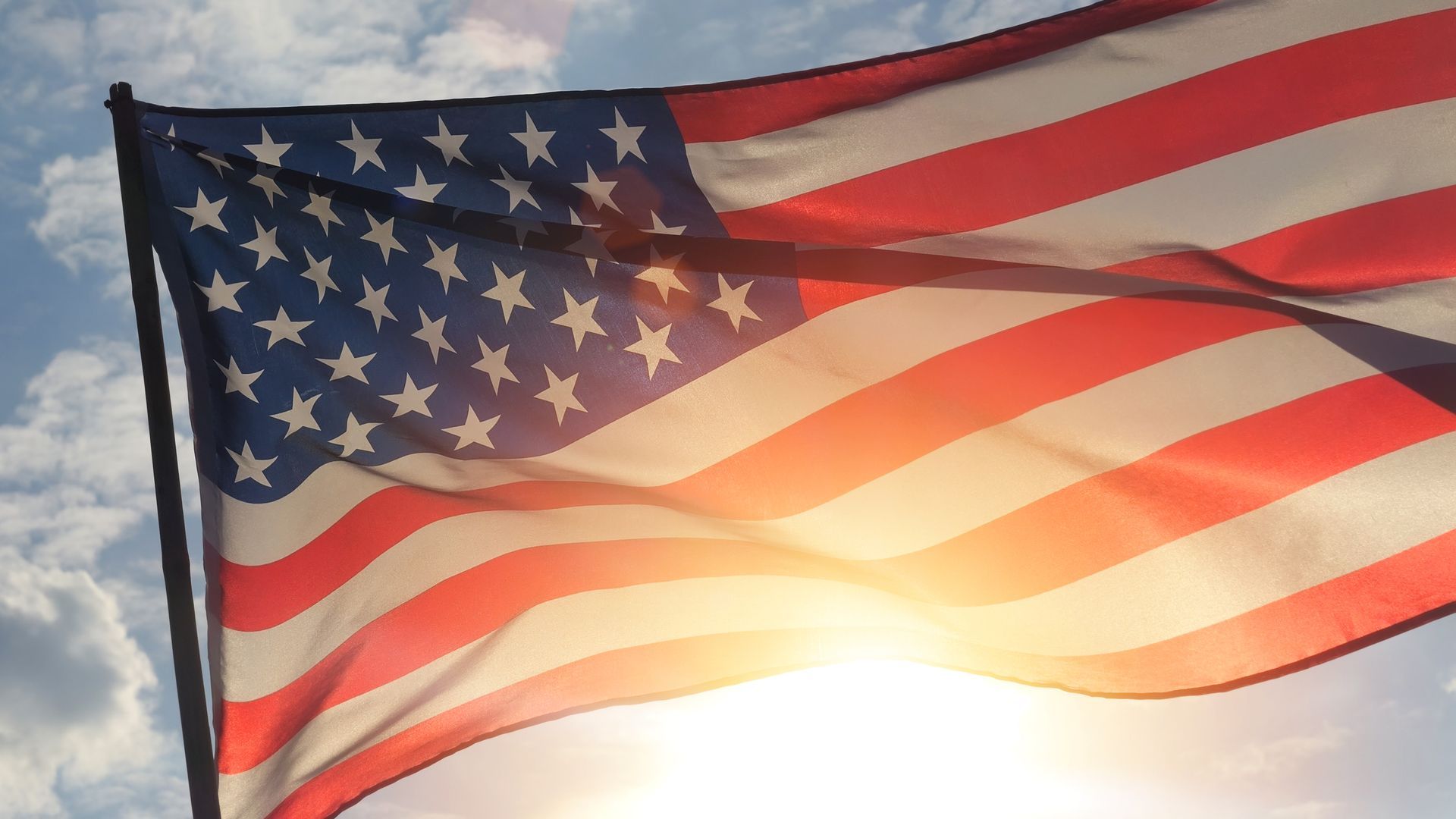 An American flag waving against a bright, sunlit sky with scattered clouds.