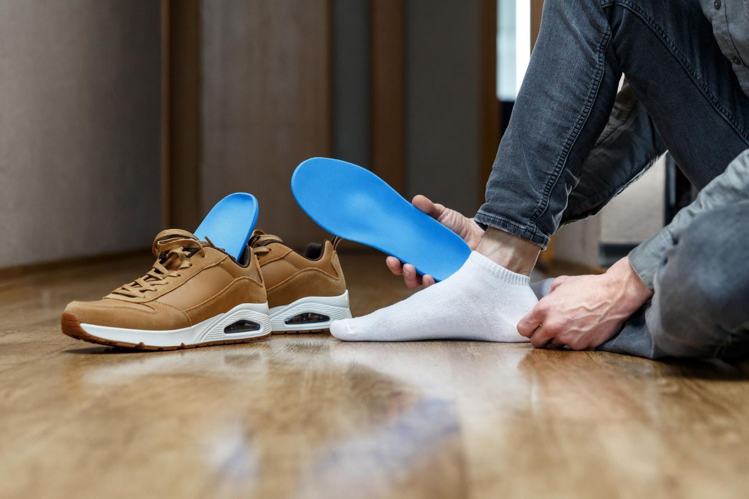 Person putting on white sock and tan sneaker with blue insole on a wooden floor.