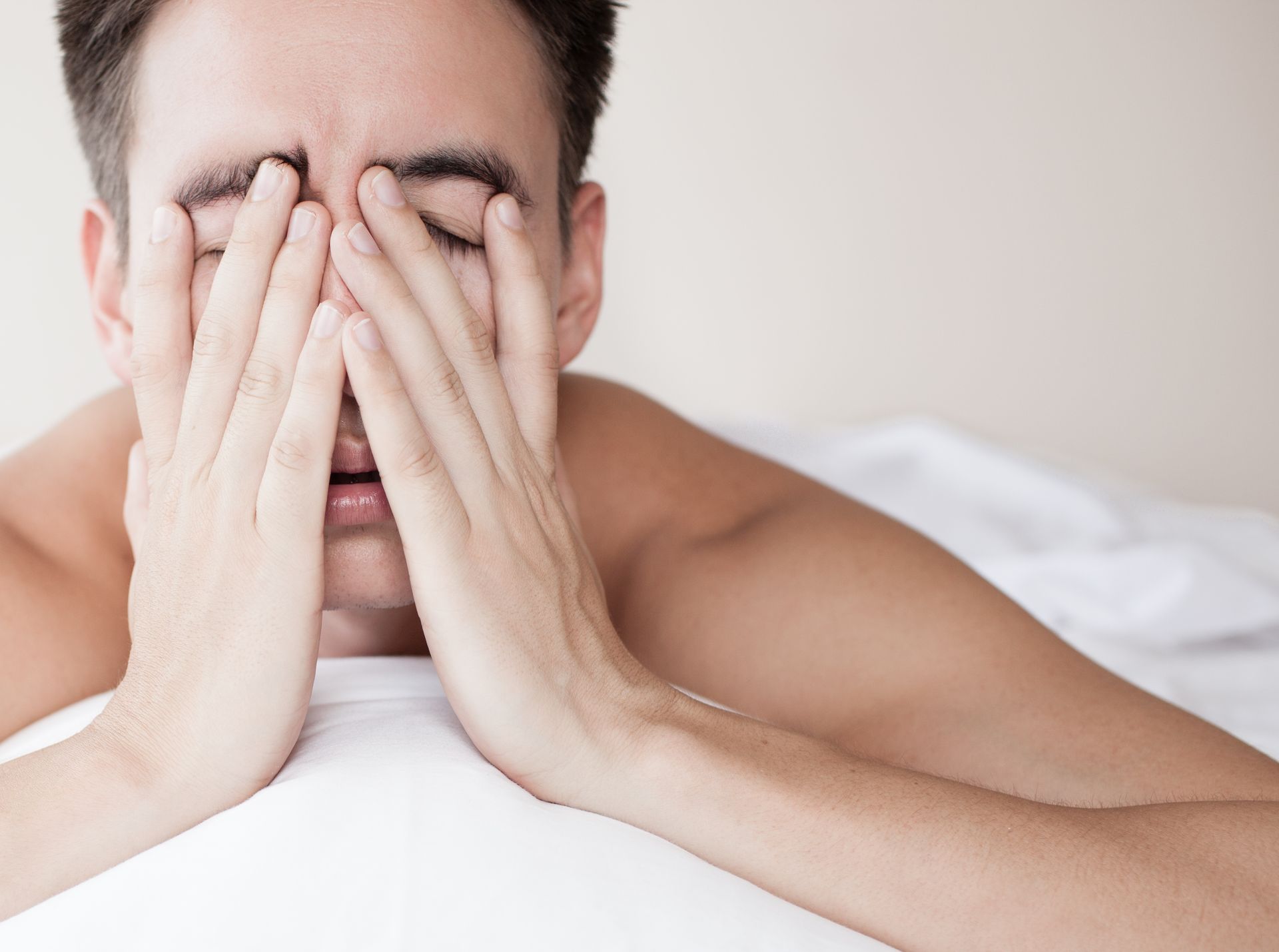 Man with hands covering his eyes, looking tired while lying on a bed.