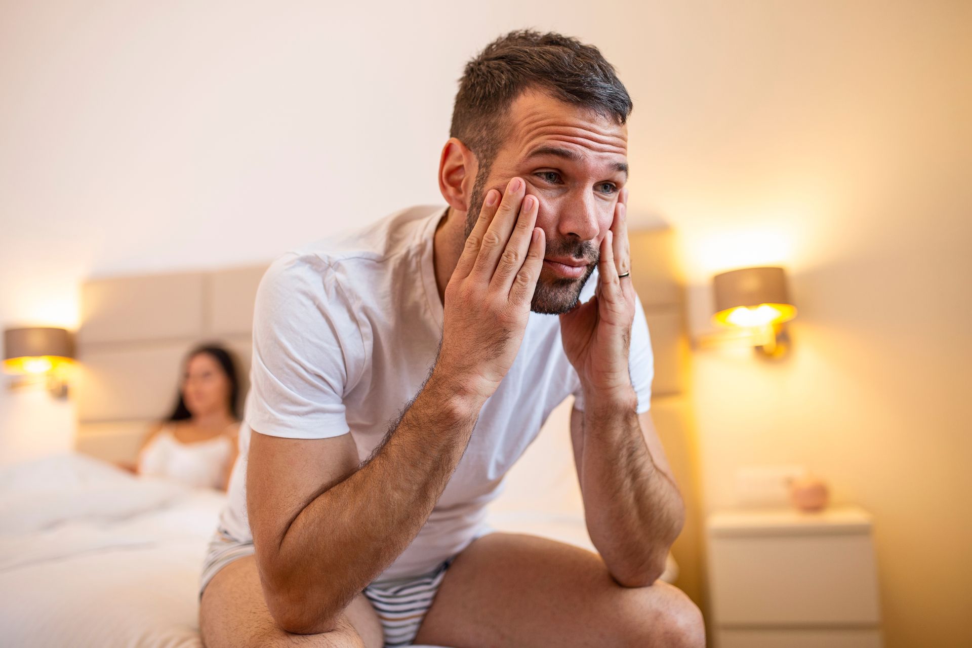 Man sits on bed, looking upset; woman in background; bedroom setting.