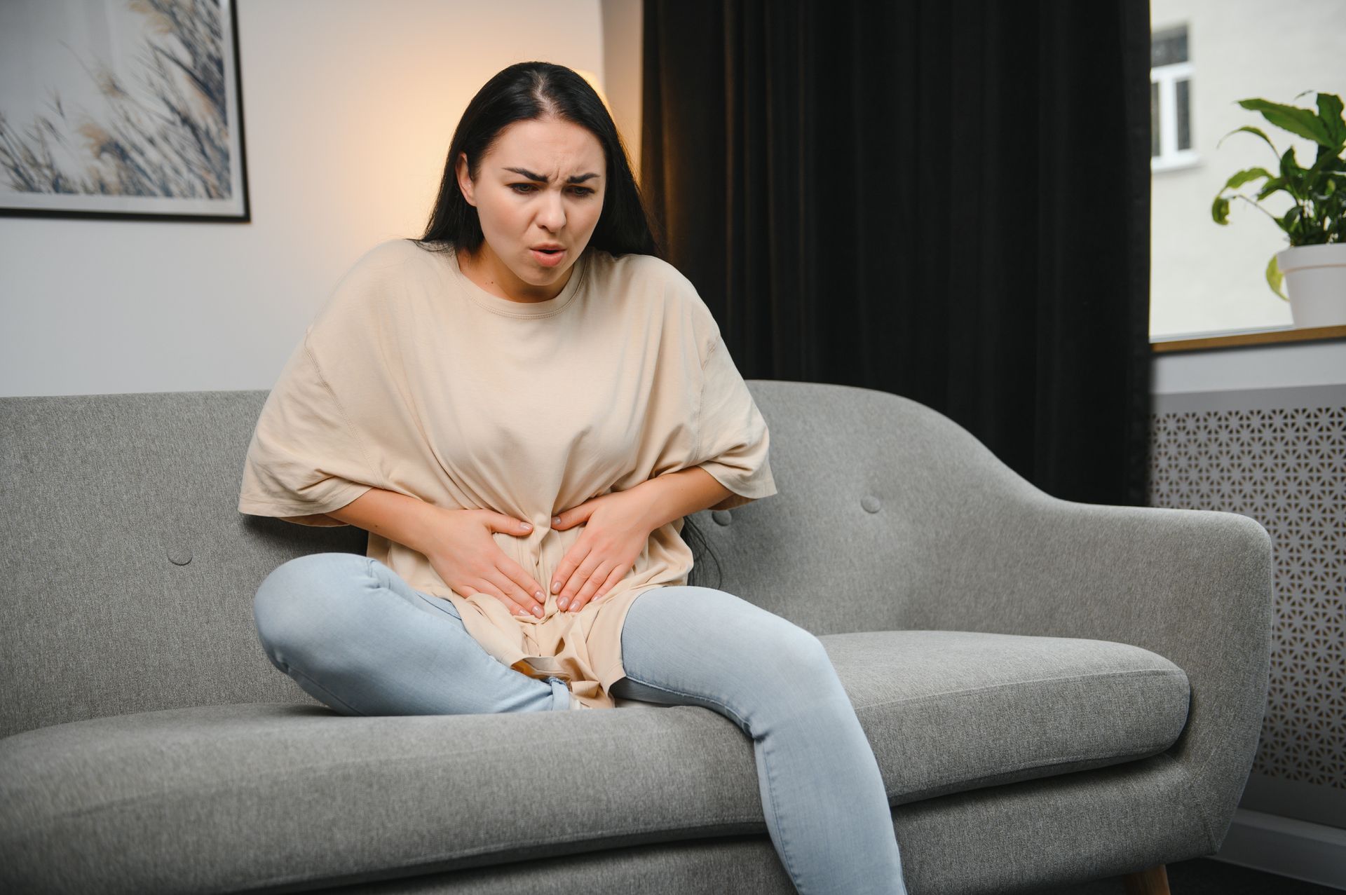 Woman sits on a couch, clutching stomach, displaying pain. Neutral-toned clothing.
