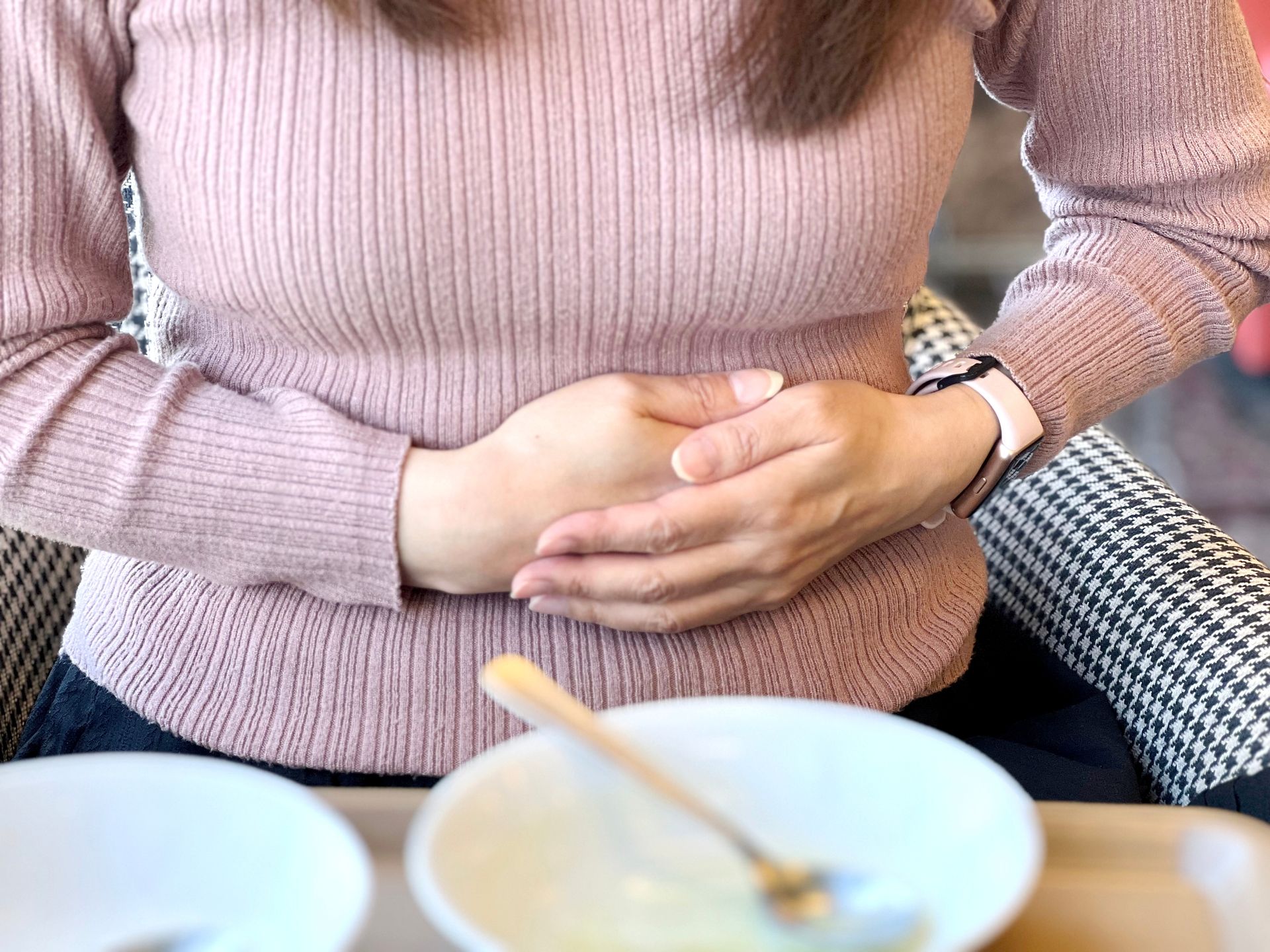 Woman in pink sweater holding stomach, possibly in pain, at a table with a bowl and spoon.