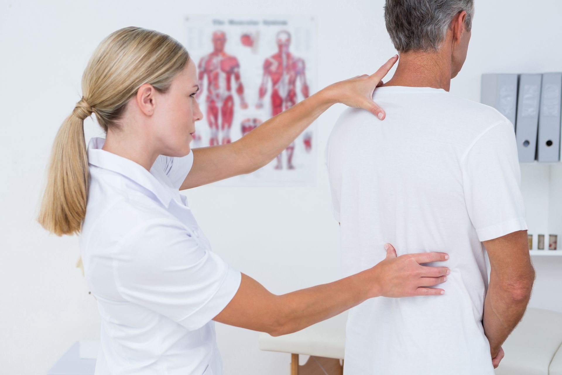 Blonde woman in white shirt examines the back of a man in a white shirt, in a medical office.