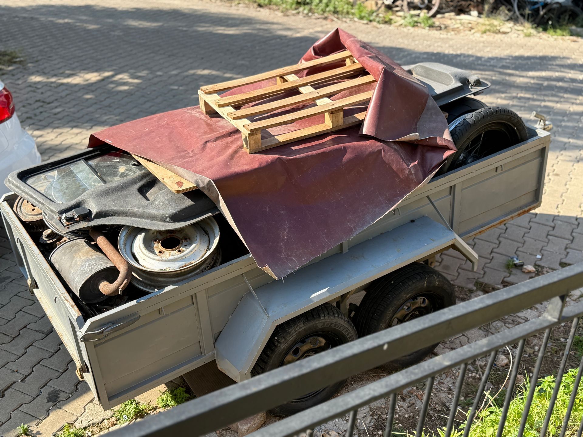 Gray trailer filled with items, covered by a maroon tarp and topped with a wooden pallet. Parked outdoors.