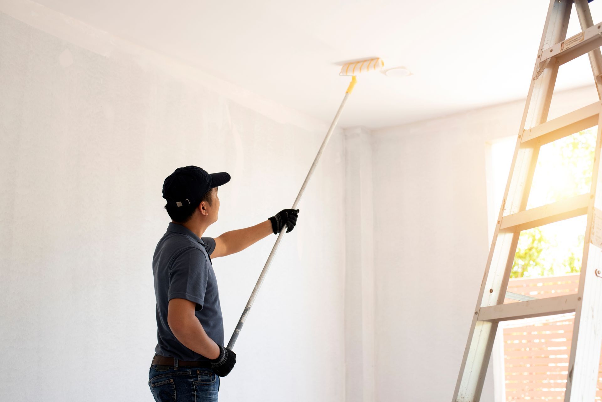 Person paints a white ceiling with a paint roller, standing near a ladder in a room with a sunny window. Person paints a white ceiling with a paint roller, standing near a ladder in a room with a sunny window.