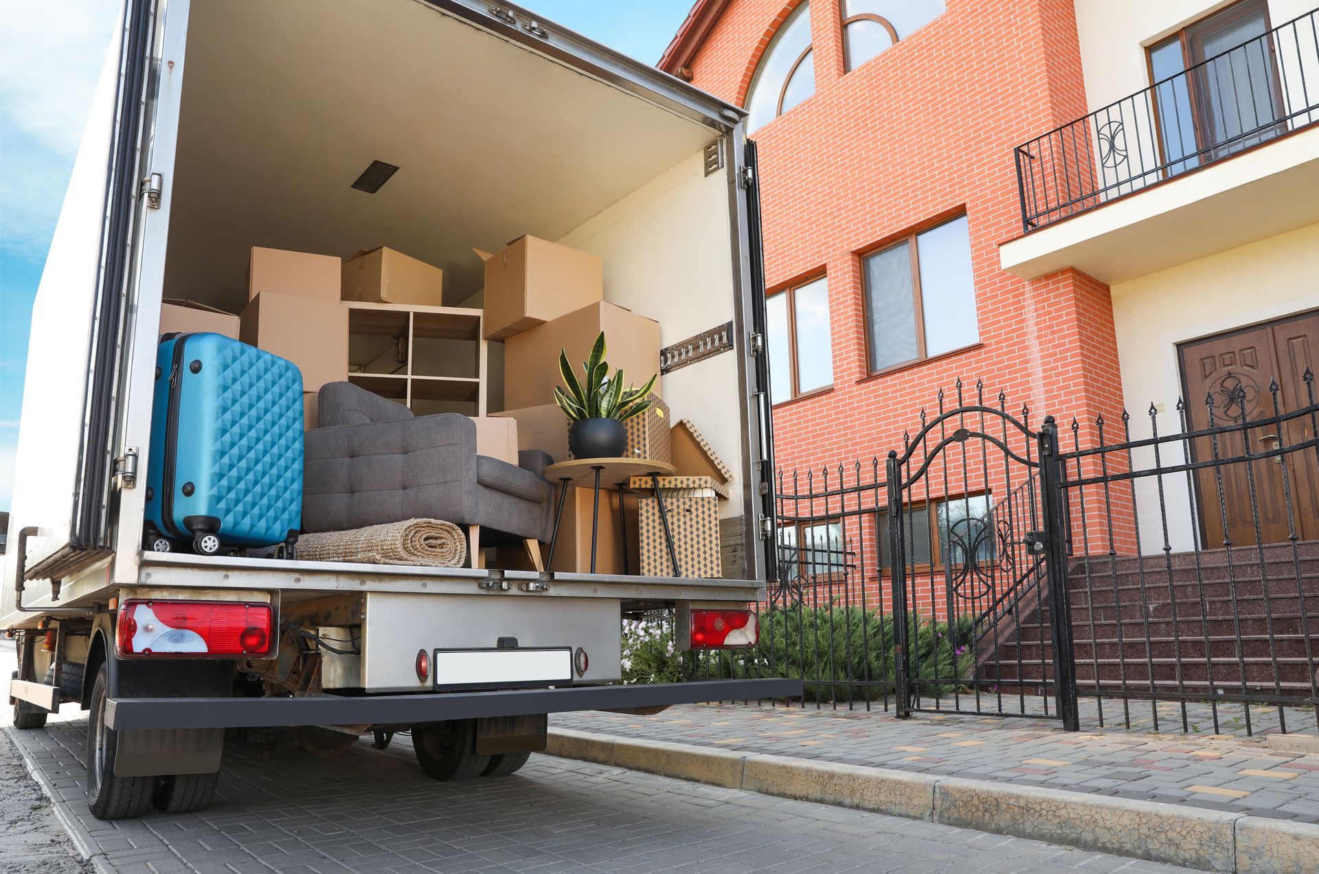 Moving truck filled with furniture and boxes, parked in front of a brick house with a wrought iron fence.