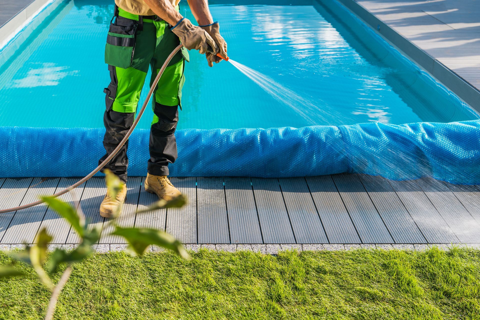 Person sprays water near a pool. They wear work clothes and gloves. Blue pool cover and green grass are visible. Person sprays water near a pool. They wear work clothes and gloves. Blue pool cover and green grass are visible.