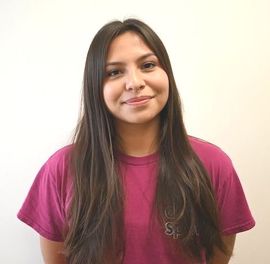 Smiling person with long dark hair wearing a magenta T-shirt against a light background