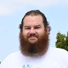Bearded man in a white shirt standing outdoors against a blue sky