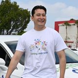 Smiling person in a white T-shirt standing beside a white car outdoors on a cloudy day