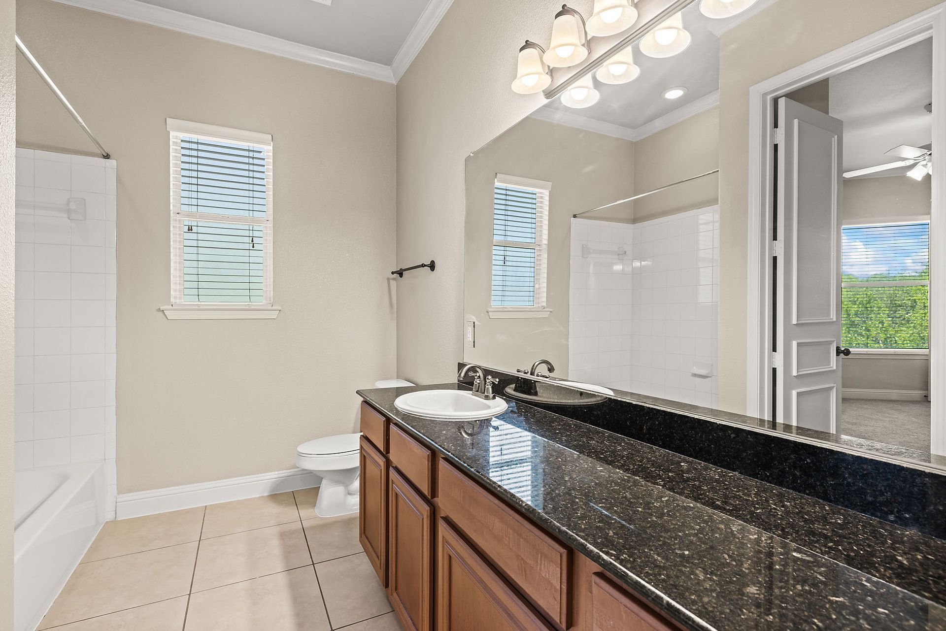 Bathroom with a long vanity, brown cabinets, black countertop, white toilet, and two small windows at Marquis at TPC, offers apartments in San Antonio, TX. A doorway leads to a room with a view of greenery.