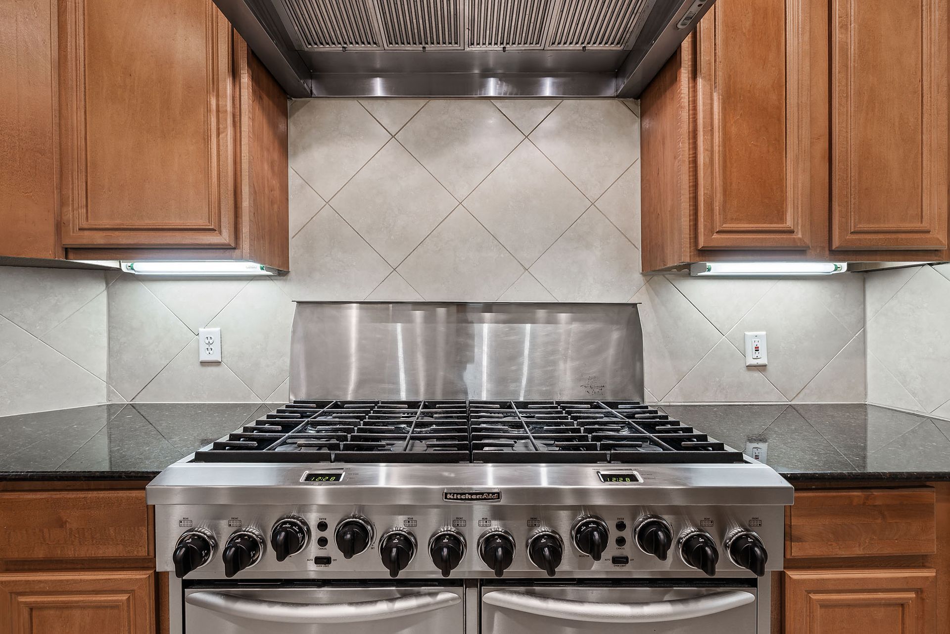 A stainless steel stove and range hood are centered between wooden cabinets. White diagonal tiles make up the backsplash.