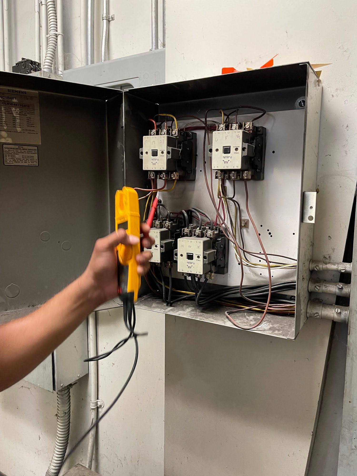 Open electrical control panel with a hand holding a yellow multimeter probe.