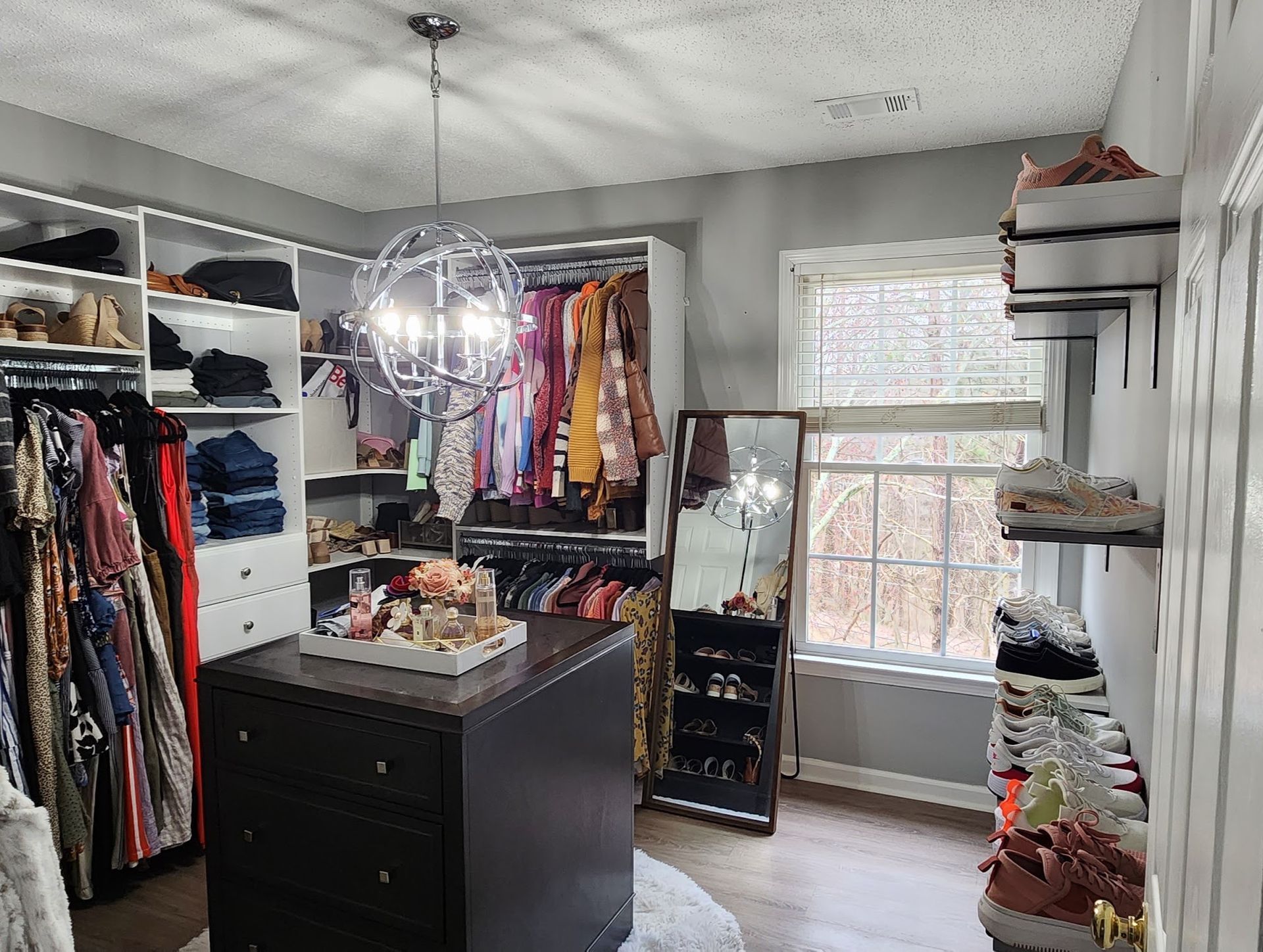 Walk-in closet with shelves, hanging clothes, dresser, mirror, window, and shoes arranged on the right.