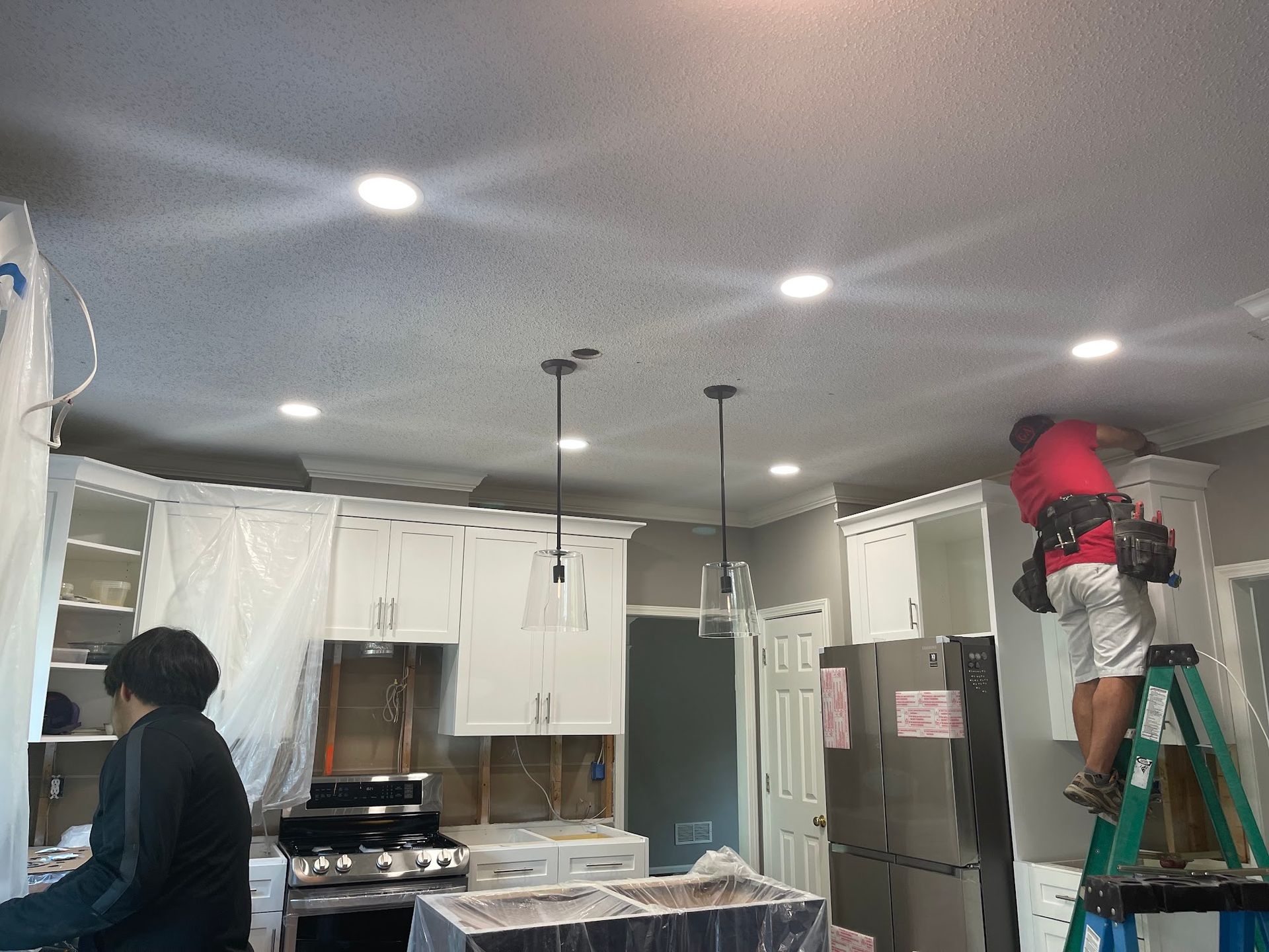 Person on ladder installing a ceiling light in a kitchen with white cabinets and stainless steel fridge