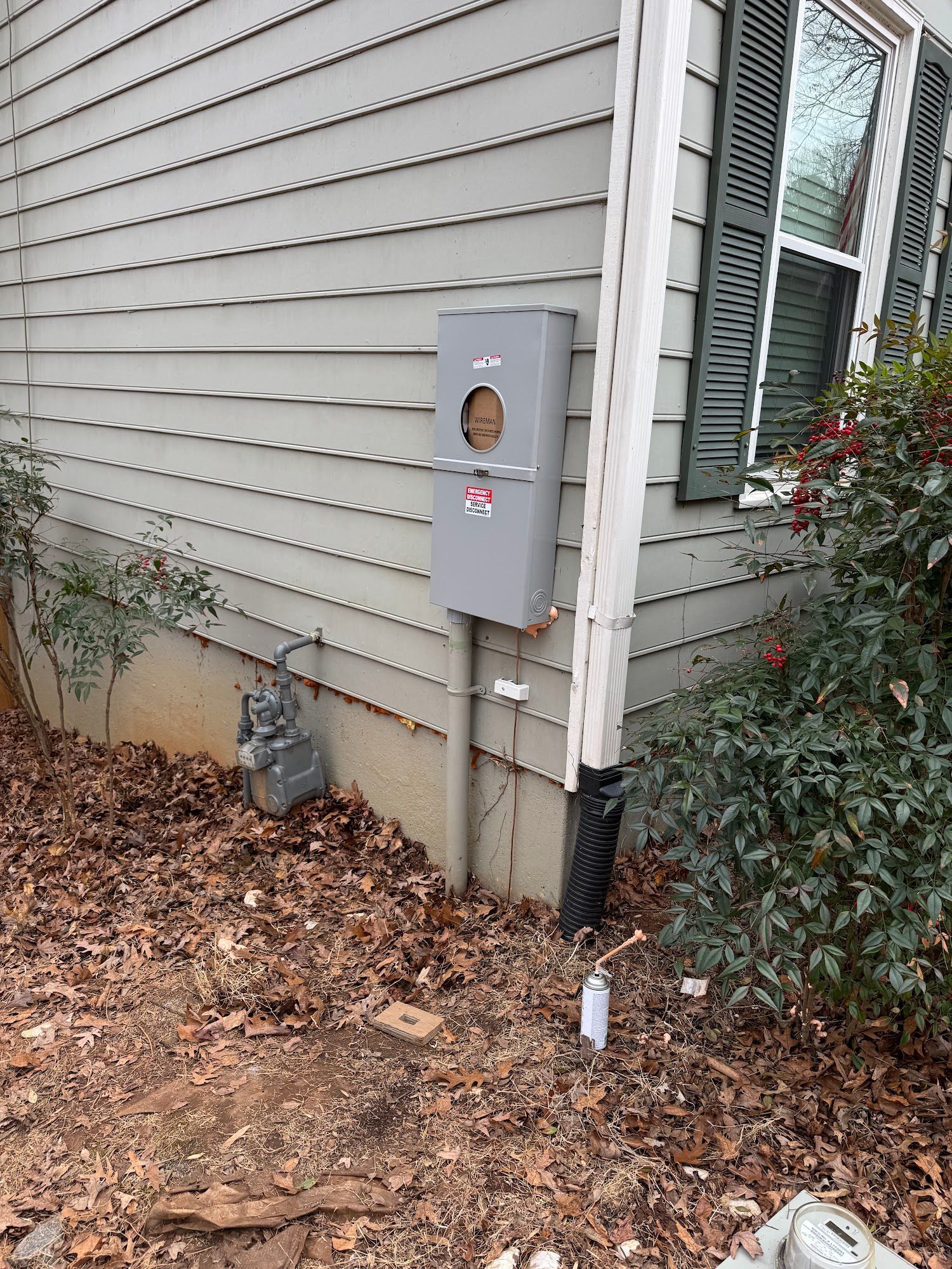 Side of a house with gray siding, utility meter box, and leaf-covered ground beside shrubs.