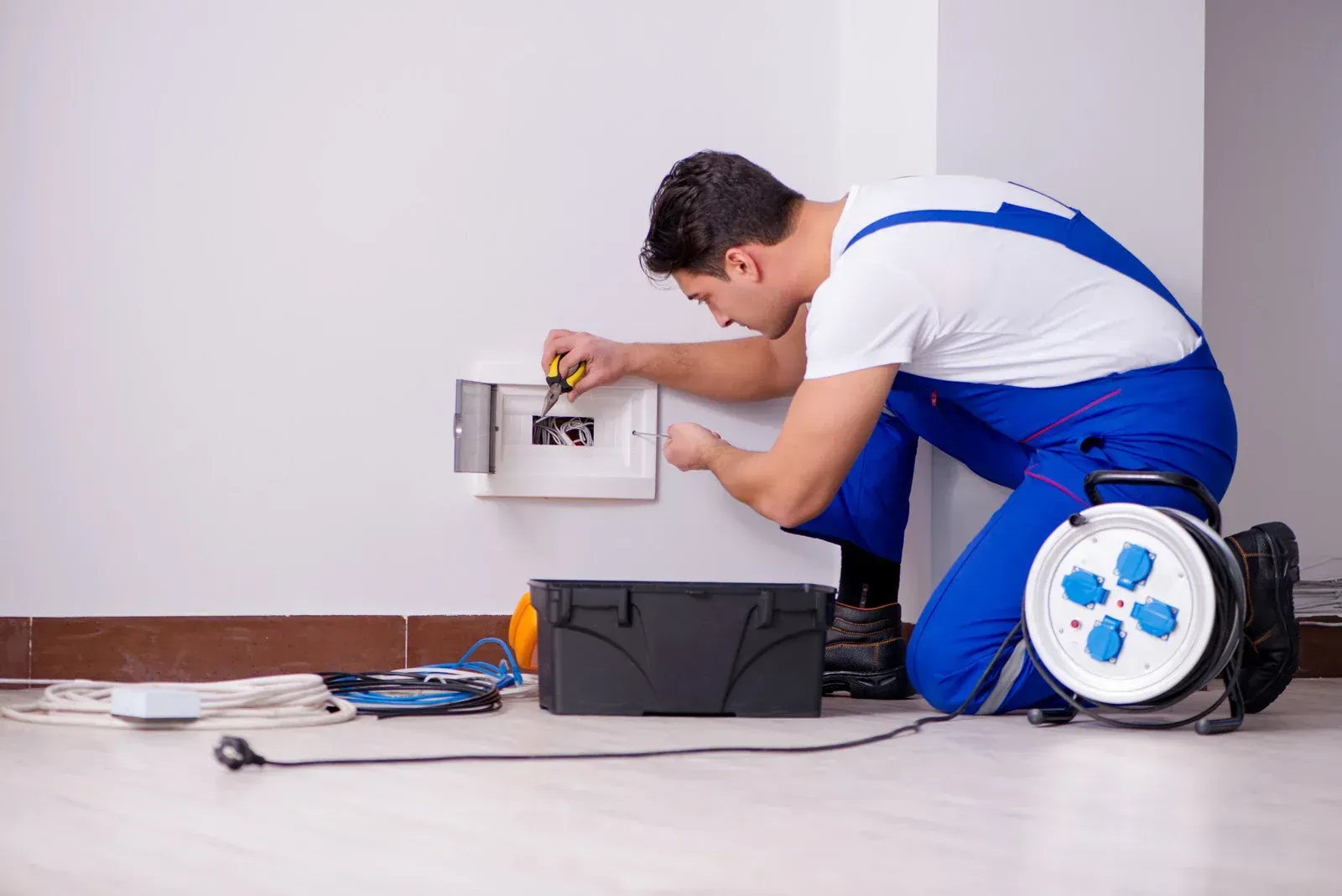 Technician in blue overalls repairs a wall outlet, with tools and cable reel on the floor.