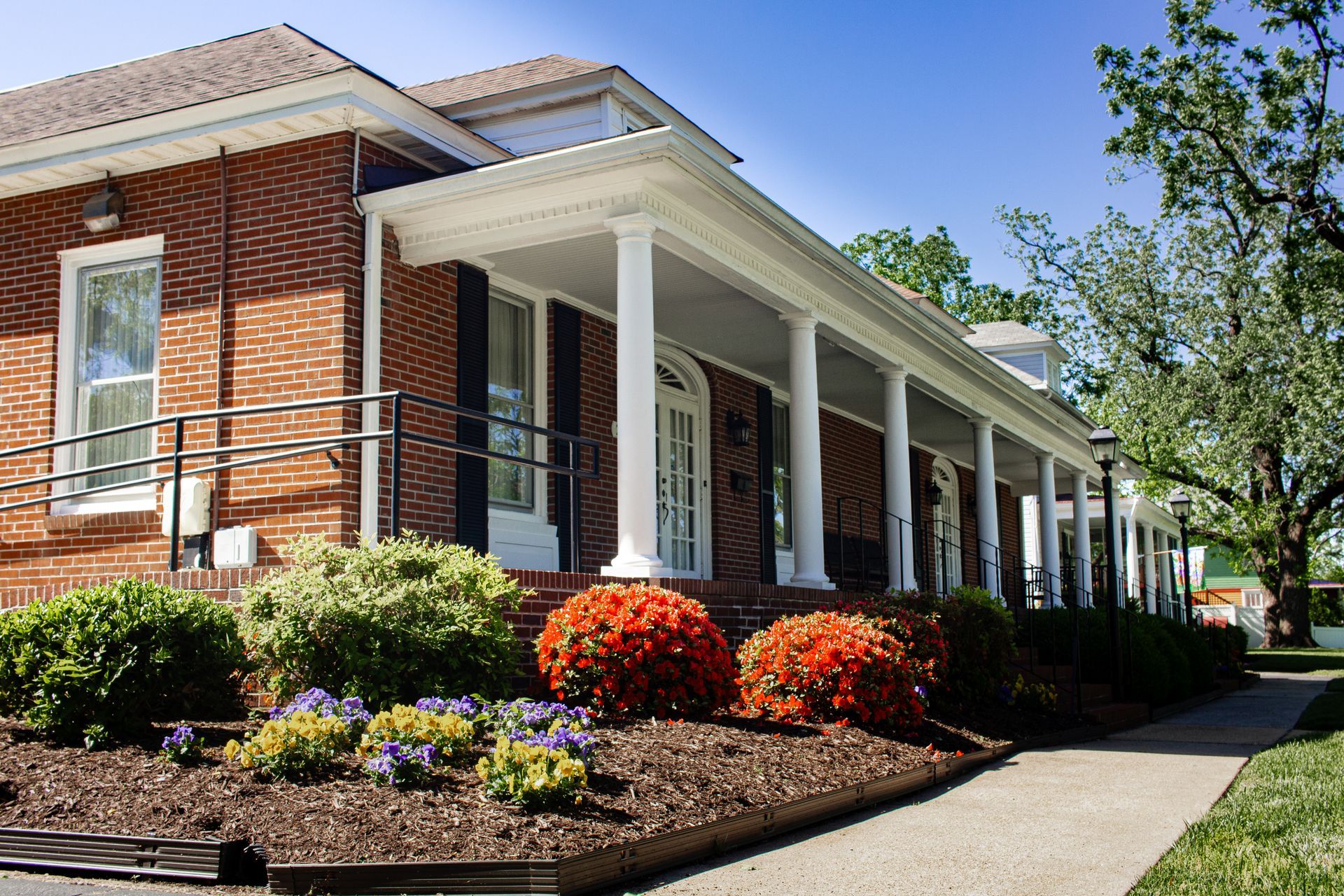 Brick building with white columns, flower beds, and a sidewalk on a sunny day