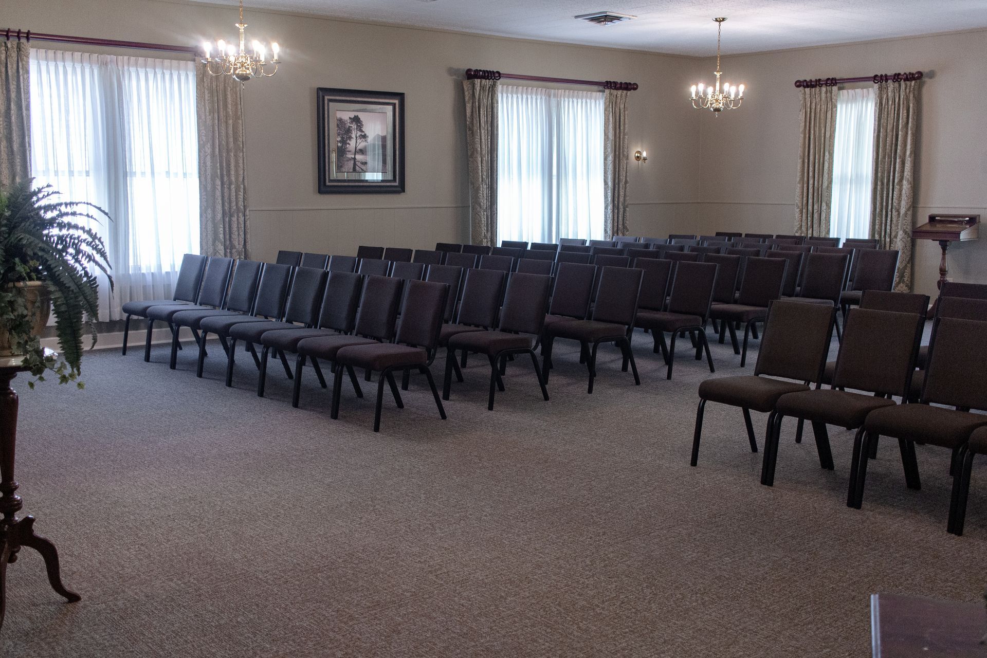 Empty meeting room with rows of dark chairs, beige carpet, and windows with white curtains
