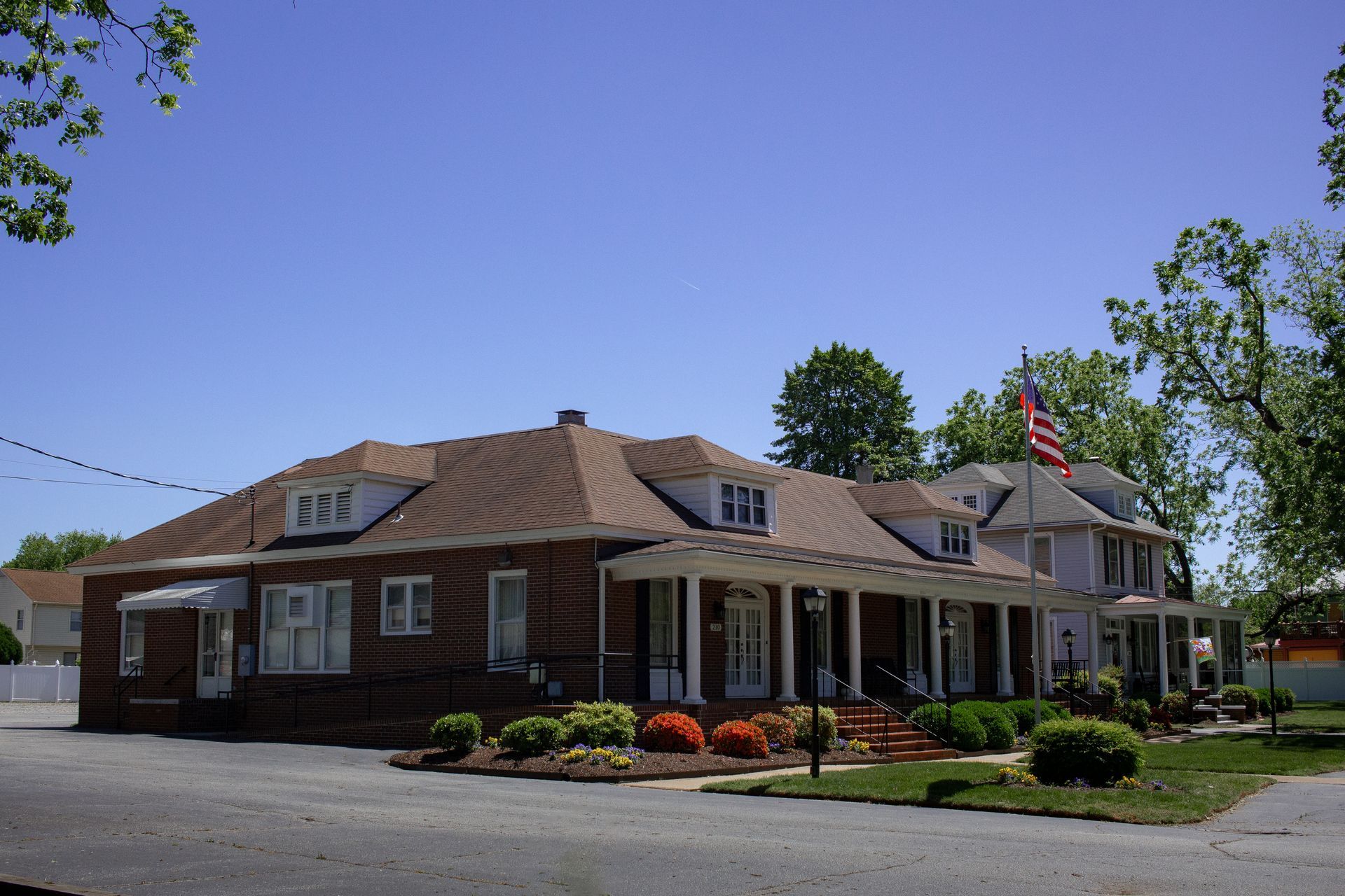 Single-story brick building with a porch, flower beds, and an American flag under a clear blue sky