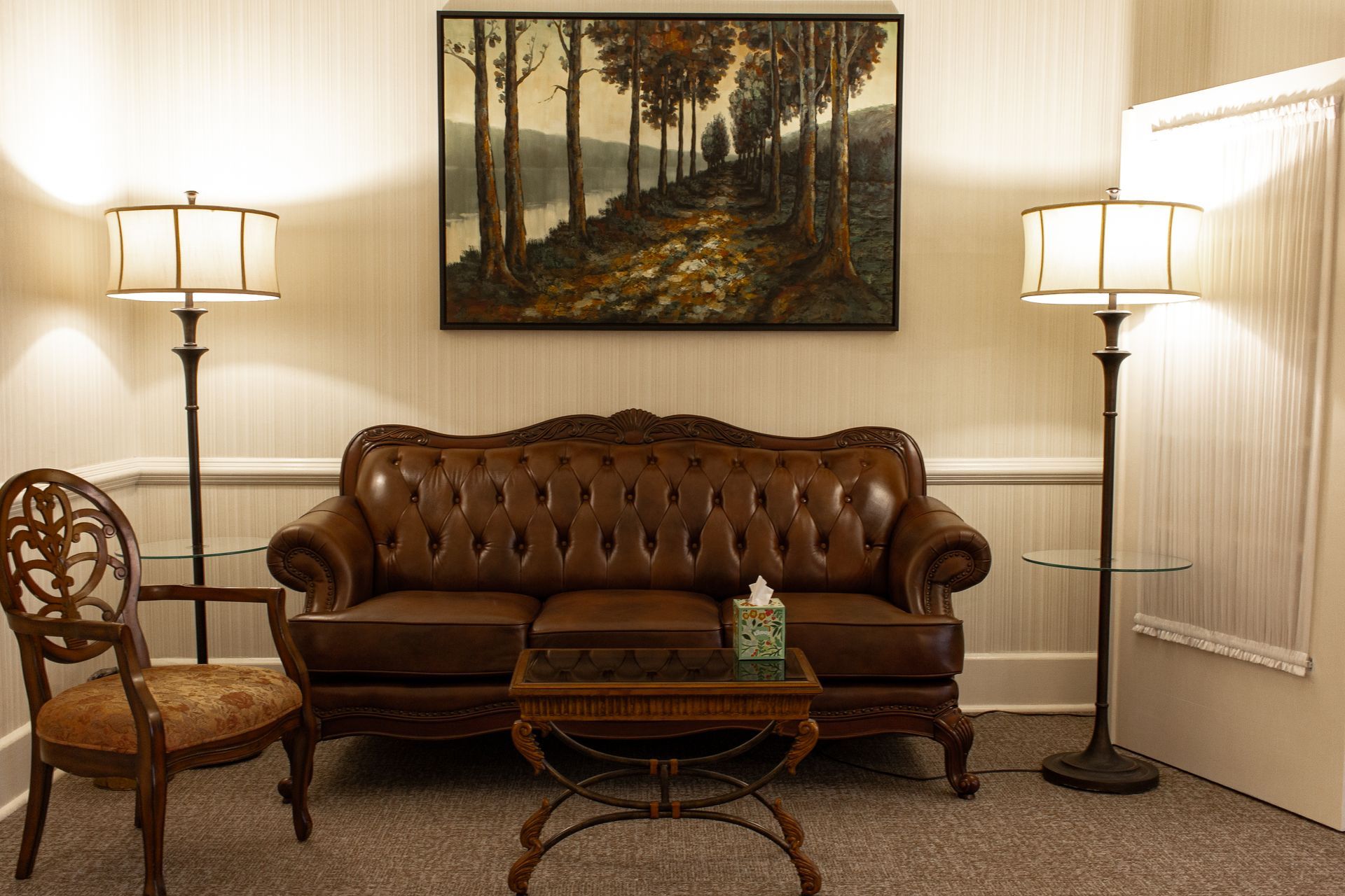 Elegant living room with leather sofa, two floor lamps, ornate chair, and landscape painting on the wall