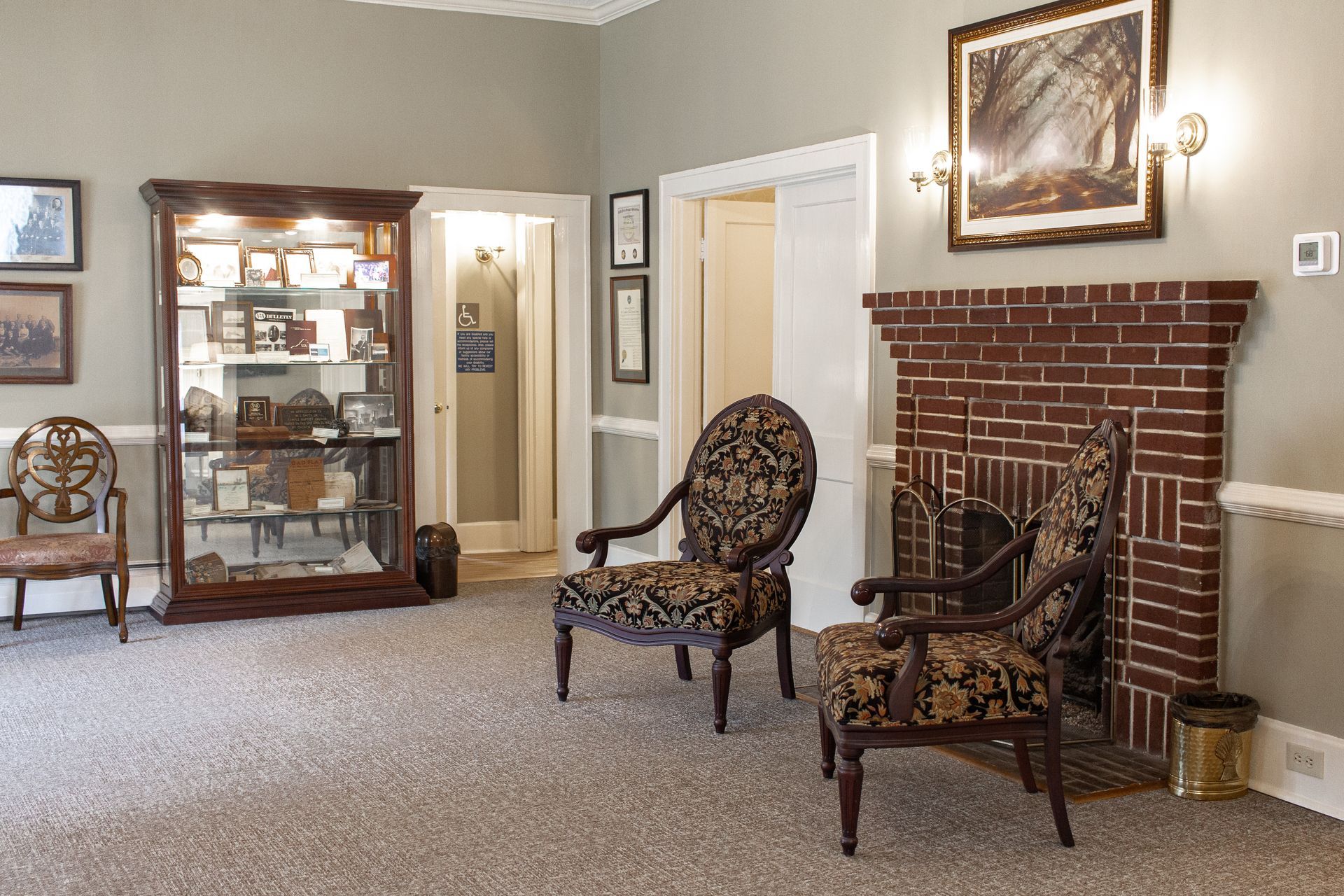 Living room with two patterned chairs, a glass display cabinet, and a brick fireplace on beige walls.