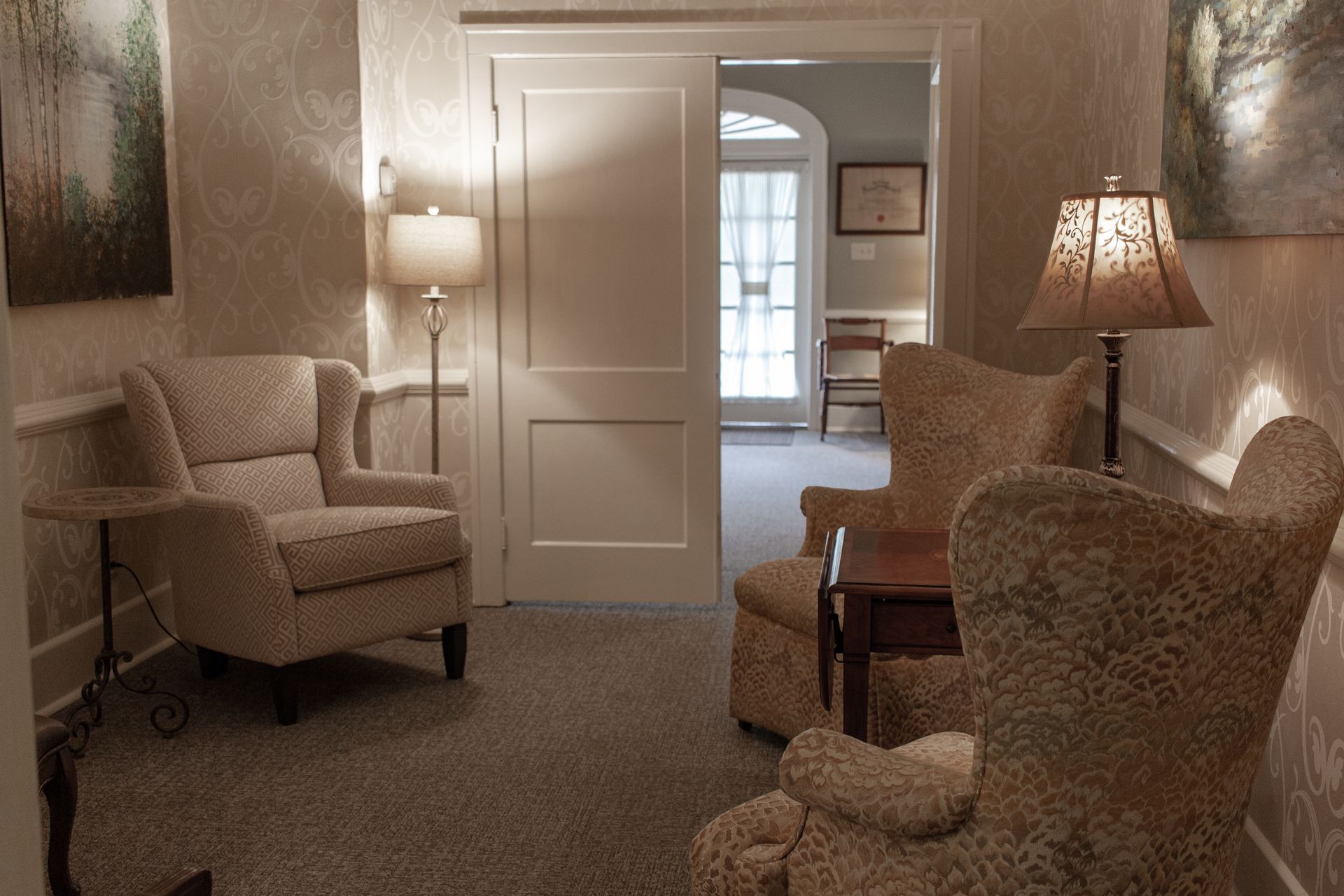 Cozy living room with beige armchairs, lamps, and an open door to a bright hallway.