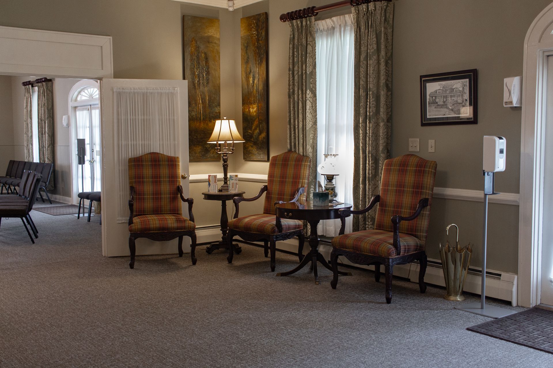 Elegant lobby seating area with plaid armchairs, table lamp, tall windows, and carpeted floor.