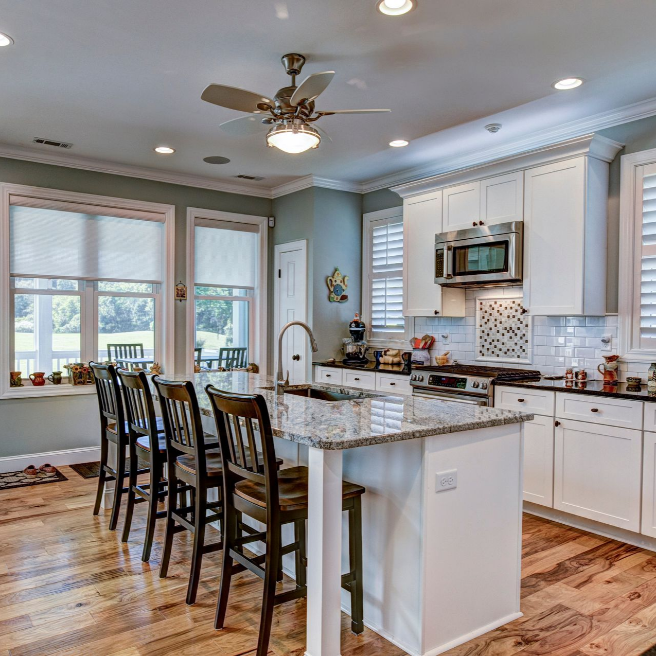 Kitchen with white cabinets, island, and wooden floor. Window with blinds, chairs, and ceiling fan. Kitchen with white cabinets, island, and wooden floor. Window with blinds, chairs, and ceiling fan.