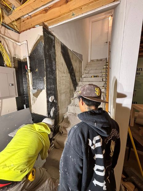 Two workers in a construction area stand near a staircase, one wearing a bright yellow hoodie and the other a dark cap.