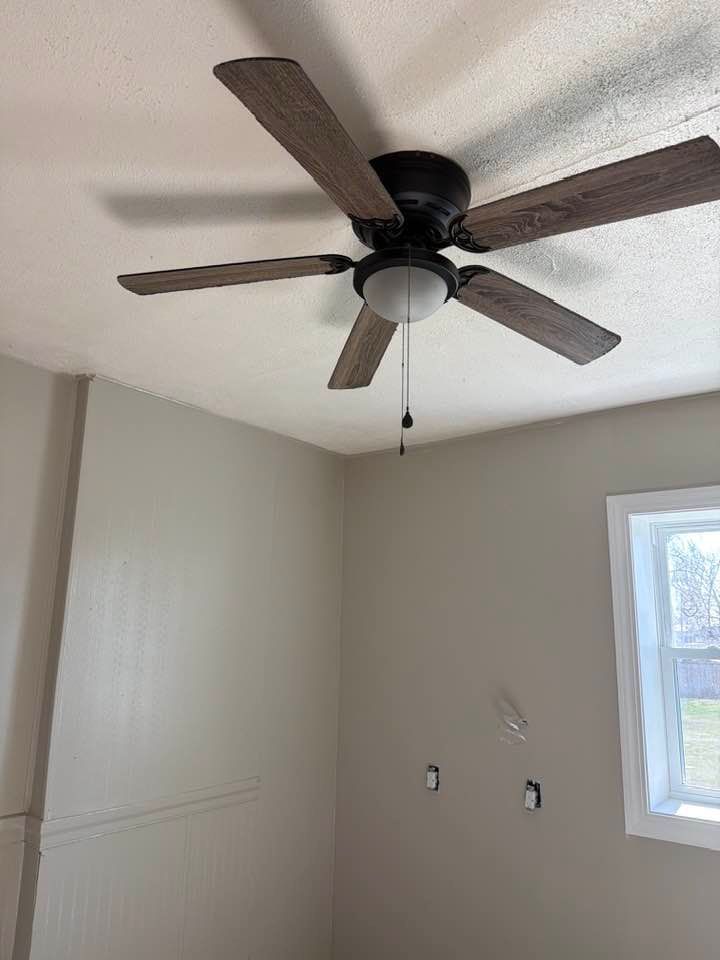 A ceiling fan with dark wood-toned blades and a center light fixture centered in a room with light grey walls and a window.