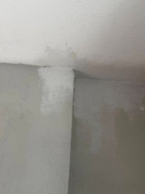 A close-up view of a textured ceiling corner showing a patch of lighter, unpainted white plaster against a gray surface.