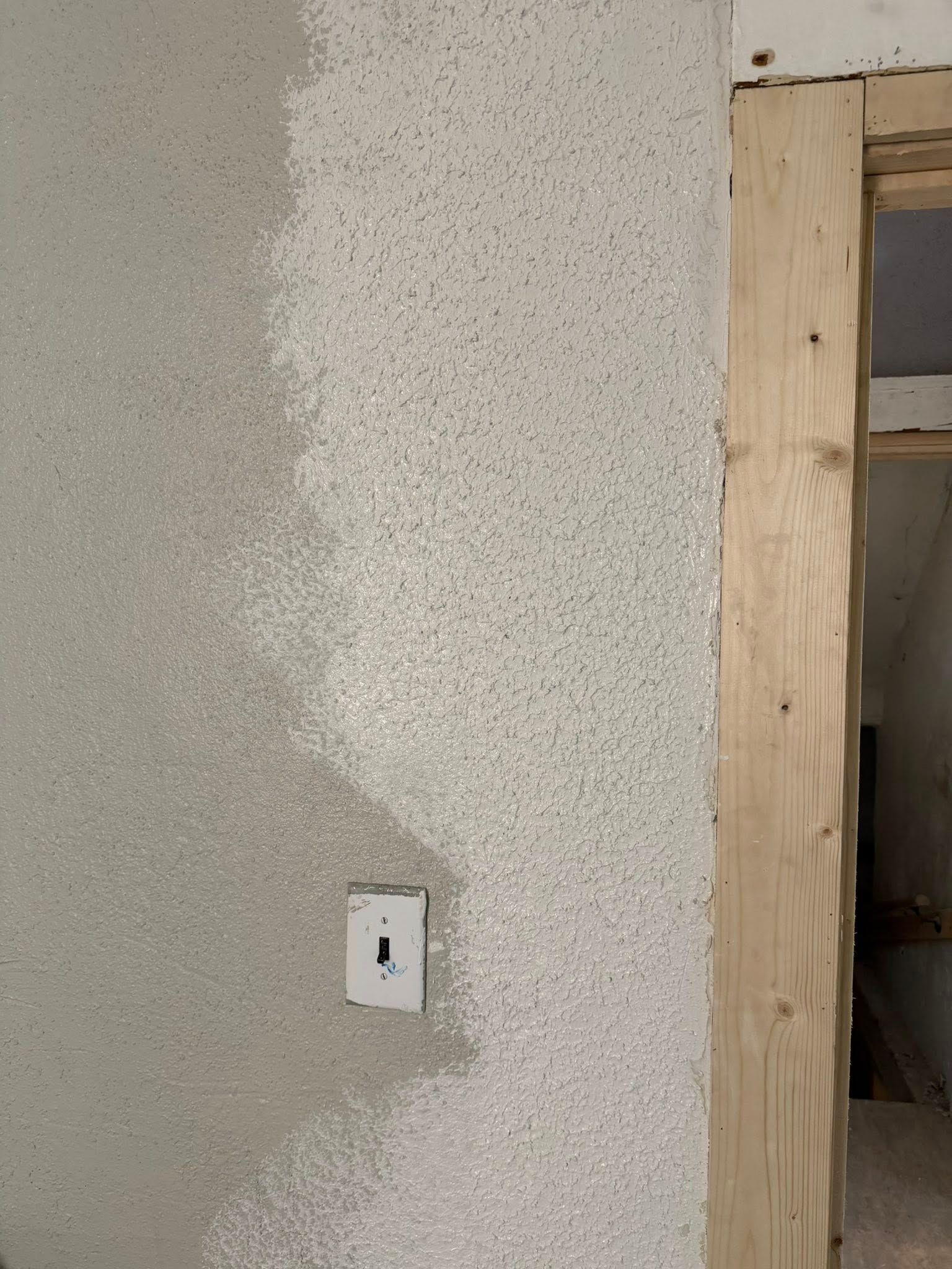 A textured, speckled gray wall is partially painted with a fresh, lighter coat next to an unfinished wooden door frame.