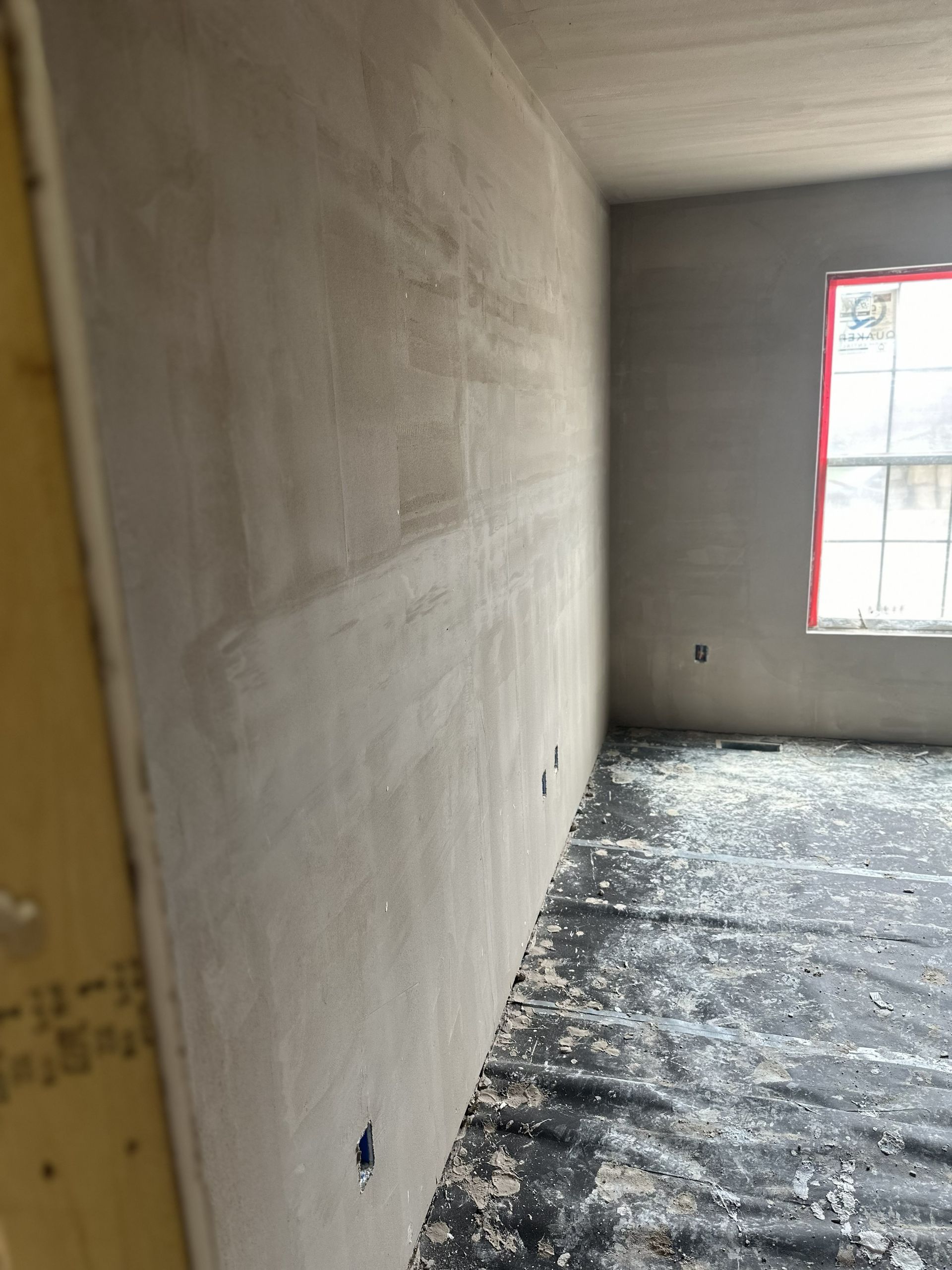 A room under construction with newly plastered gray walls, a window with a red frame, and a floor covered in debris.