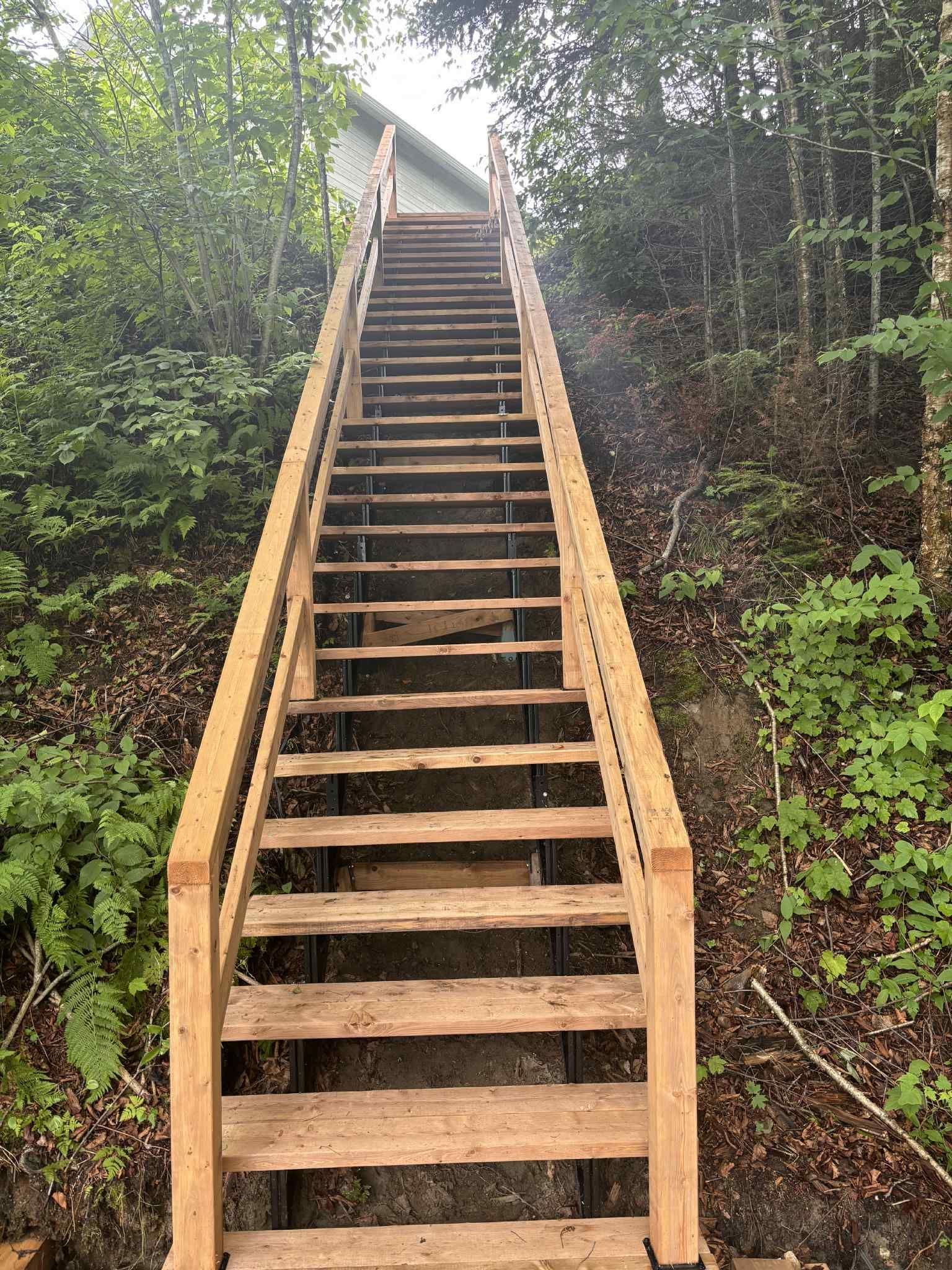 Un escalier droit et raide en bois, muni de rampes, menant à une maison sur une colline boisée.