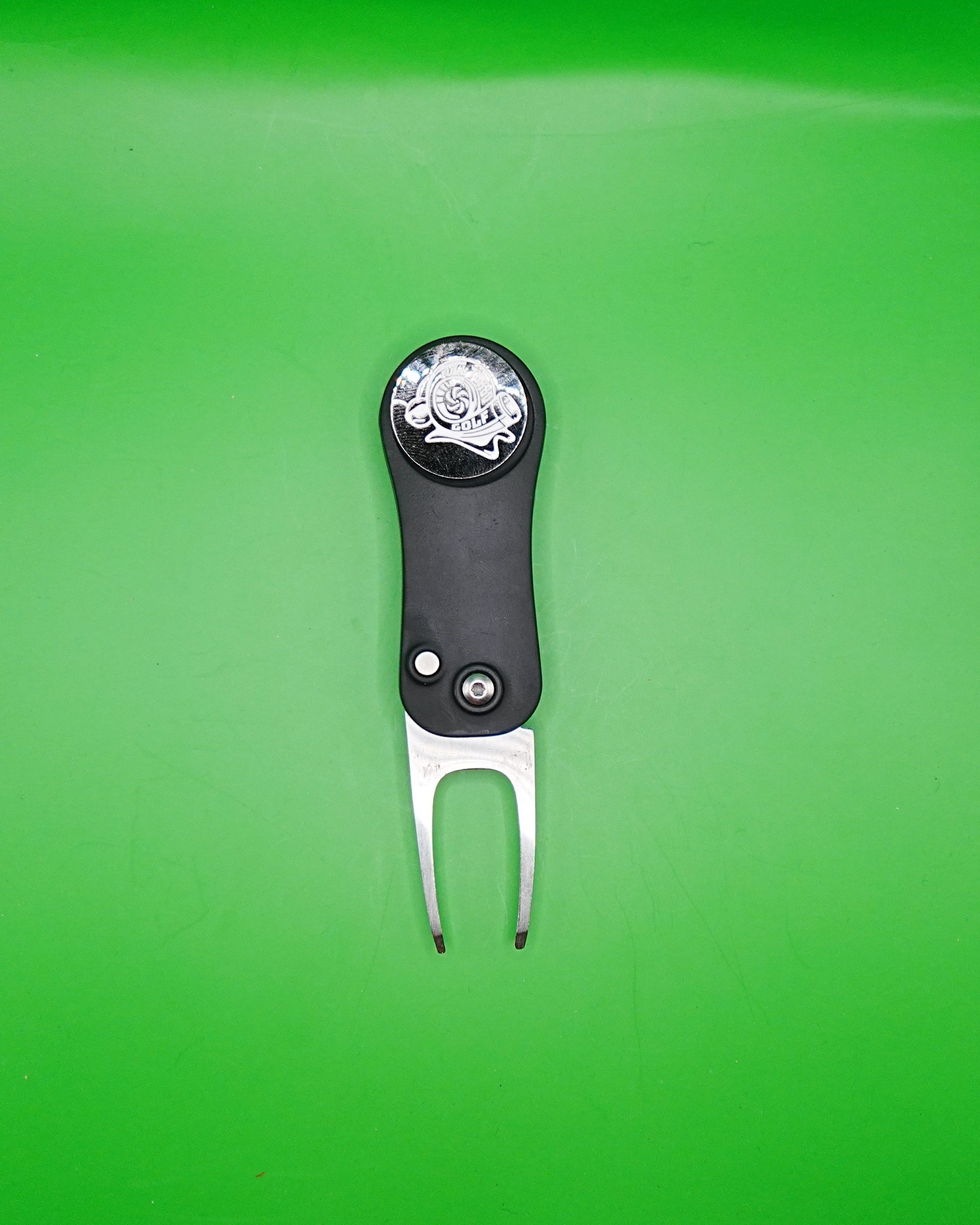 Black golf divot tool with silver prongs on green background.