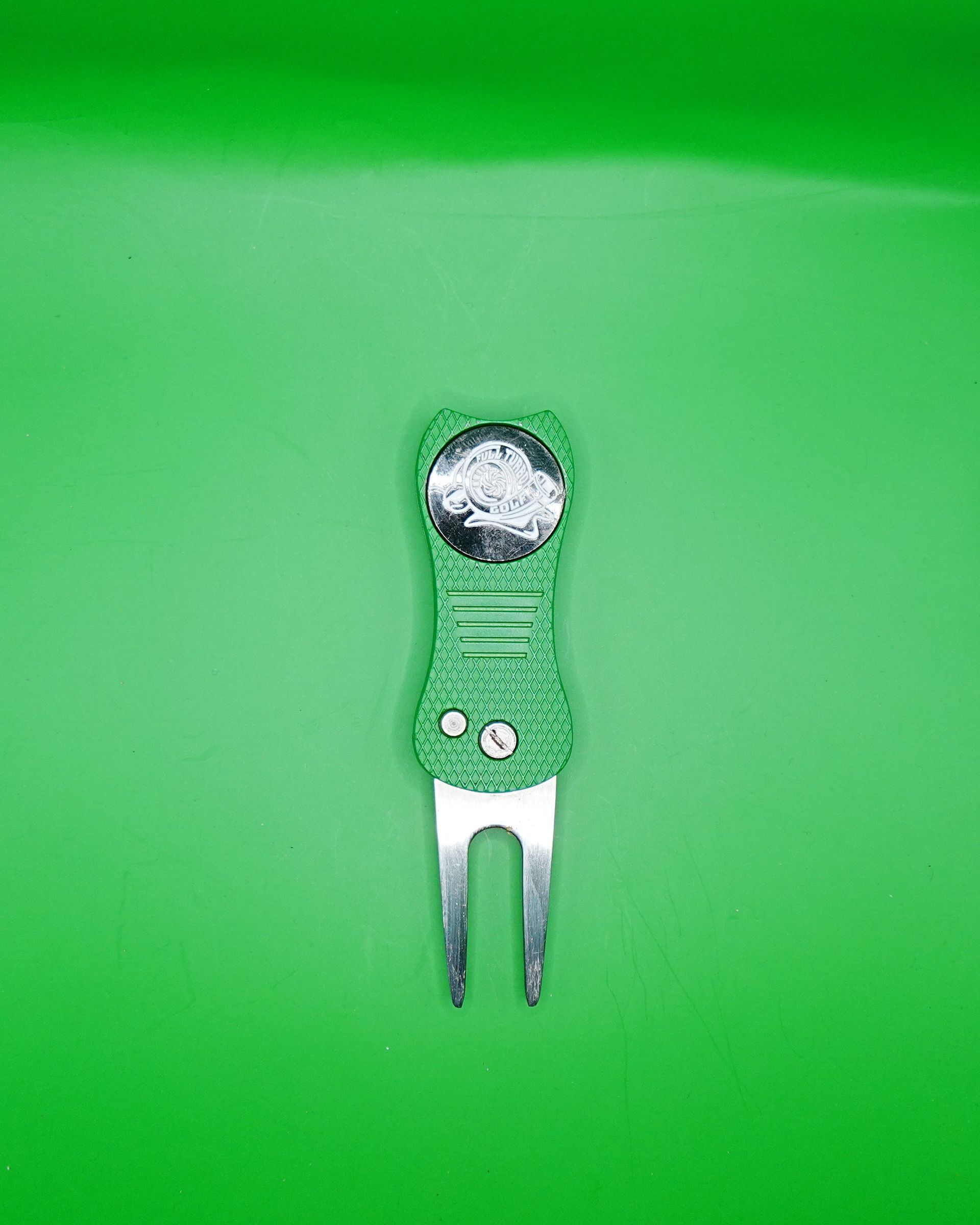 Green golf divot repair tool with a metal fork on a green background.