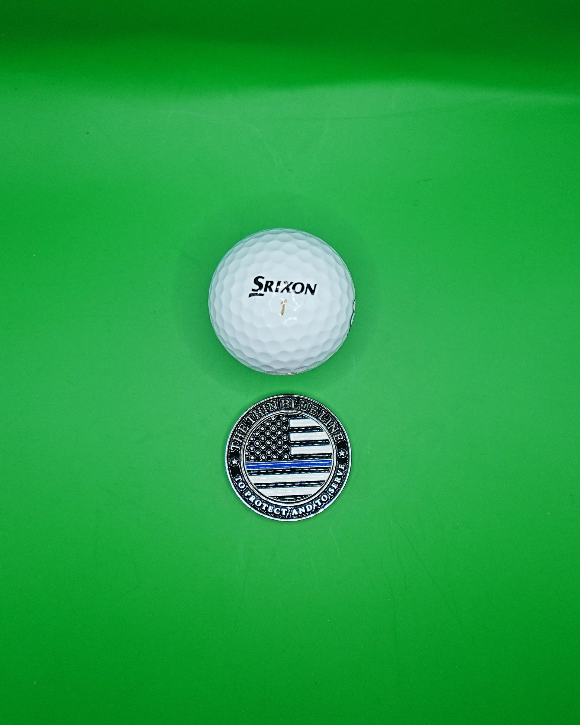 Golf ball and Thin Blue Line coin on green surface.
