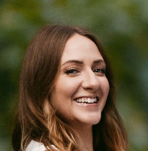 Woman with long brown hair smiles broadly, outdoors, against a blurred green background.